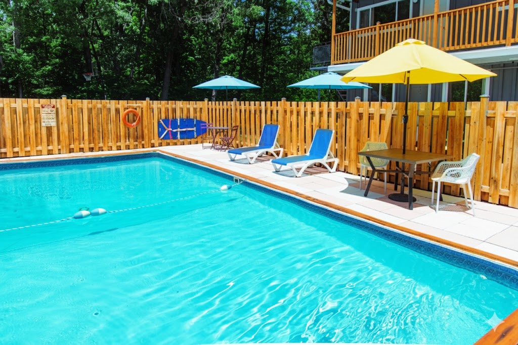 Discover the charming outdoor pool area at Whispering Pines Motel Grand Bend Pool 1. Dive into turquoise waters, relax on sun loungers, or enjoy a shaded table under umbrellas. A wooden fence surrounds you as trees provide a lush, green backdrop for your perfect getaway.